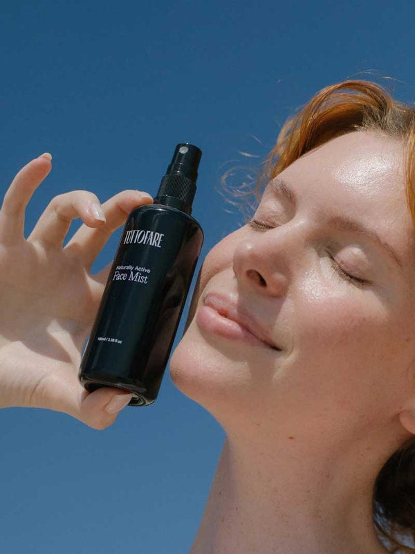 A person with fair skin and reddish hair smiles with closed eyes, holding a black bottle labeled Tuttofare Naturally Active Face Mist 100ml near their face against a blue sky background.