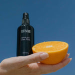 A hand holds a black bottle labeled Tuttofare Naturally Active Face Mist 100ml and half of a fresh orange against a clear blue sky.