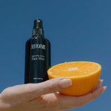 A hand holds a black bottle labeled Tuttofare Naturally Active Face Mist 100ml and half of a fresh orange against a clear blue sky.