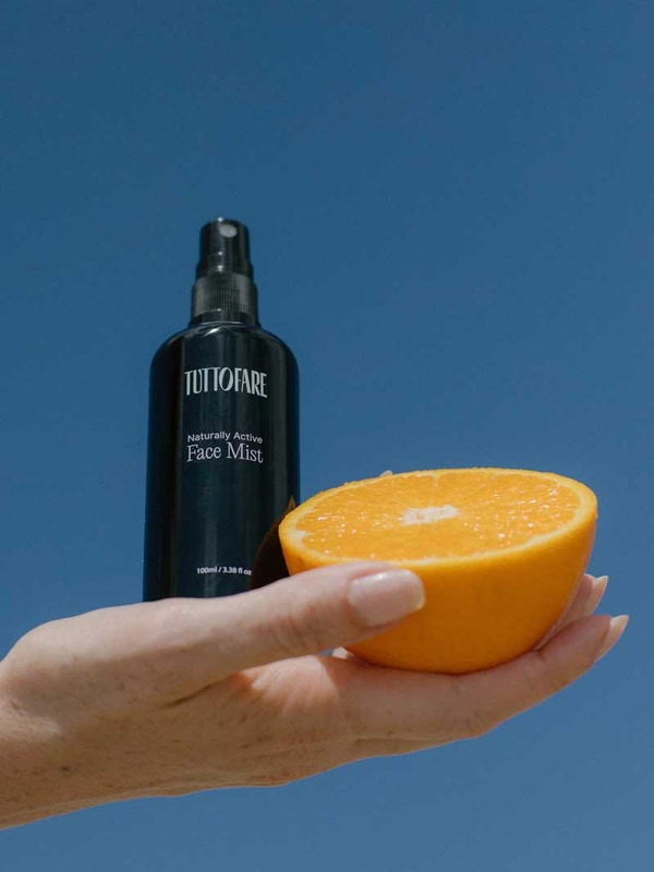 A hand holds a black bottle labeled Tuttofare Naturally Active Face Mist 100ml and half of a fresh orange against a clear blue sky.