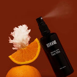 A black Tuttofare Naturally Active Face Mist 100ml spray bottle leans against a sliced orange topped with a white flower on a brown background. This hydrating mist features Snow Mushroom for a refreshing botanical boost.