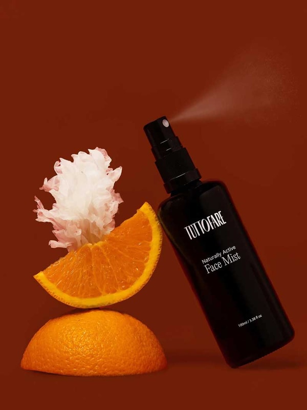 A black Tuttofare Naturally Active Face Mist 100ml spray bottle leans against a sliced orange topped with a white flower on a brown background. This hydrating mist features Snow Mushroom for a refreshing botanical boost.