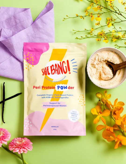 A bag of She BANG! Peri Protein POWder with KSM-66® Ashwagandha - Vanilla 420g rests on a light green surface, next to a bowl of powder with a wooden spoon, yellow and pink flowers, and a folded lavender cloth.
