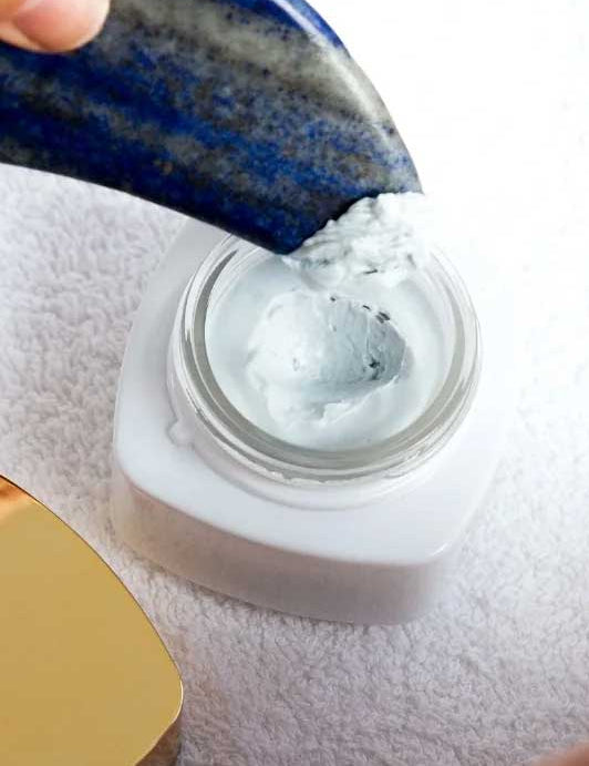 A close-up of a blue stone spatula scooping Vanessa Megan AEON Azure Regenerative Cream 50ml from an open white jar on a textured surface, with a gold-colored lid partially visible beside the jar.