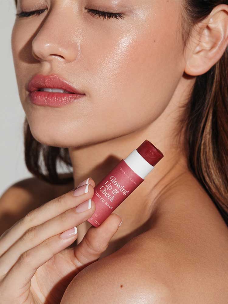 A woman with glowing skin and closed eyes holds Vanessa Megan Glowing Lip & Cheek Tinted Balm 10g, a rosy pink multi-use balm, near her face, accentuating her natural makeup look.