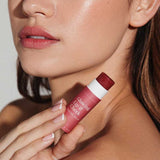 A woman with glowing skin and closed eyes holds Vanessa Megan Glowing Lip & Cheek Tinted Balm 10g, a rosy pink multi-use balm, near her face, accentuating her natural makeup look.