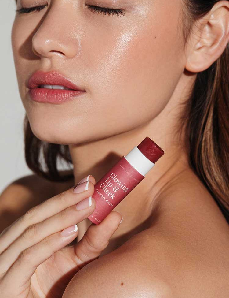 A woman with glowing skin and closed eyes holds Vanessa Megan Glowing Lip & Cheek Tinted Balm 10g, a rosy pink multi-use balm, near her face, accentuating her natural makeup look.