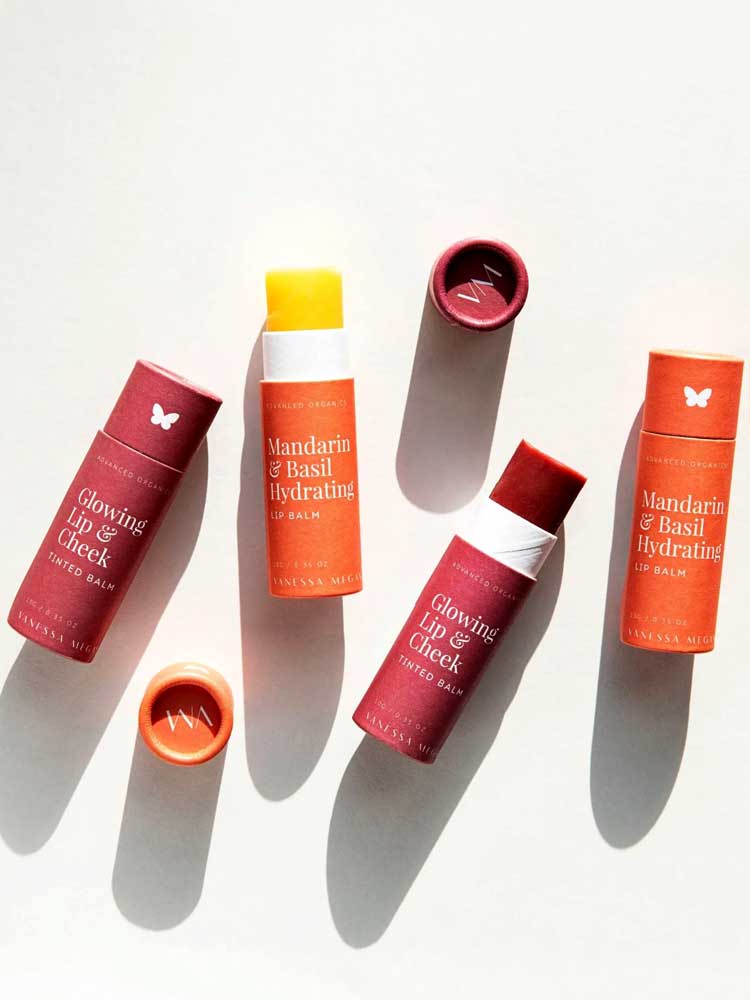 Four cylindrical tubes of tinted balm in orange and red shades, all labeled “Vanessa Megan Glowing Lip & Cheek Tinted Balm 10g,” are arranged on a white surface.
