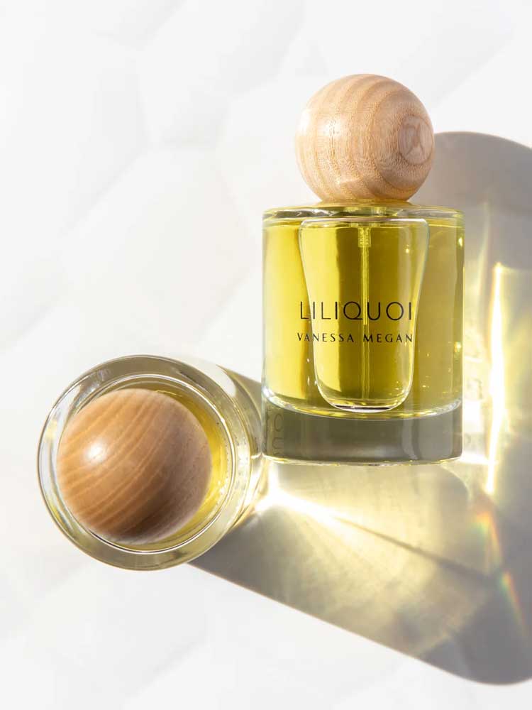 A bottle of Vanessa Megan Liliquoi 100% Natural Eau de Parfum 50ml with a wooden cap stands upright while another lies on its side, both casting soft shadows on a white surface.