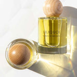 A bottle of Vanessa Megan Liliquoi 100% Natural Eau de Parfum 50ml with a wooden cap stands upright while another lies on its side, both casting soft shadows on a white surface.