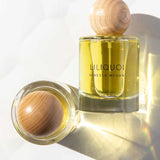 A bottle of Vanessa Megan Liliquoi 100% Natural Eau de Parfum 50ml with a wooden cap stands upright while another lies on its side, both casting soft shadows on a white surface.