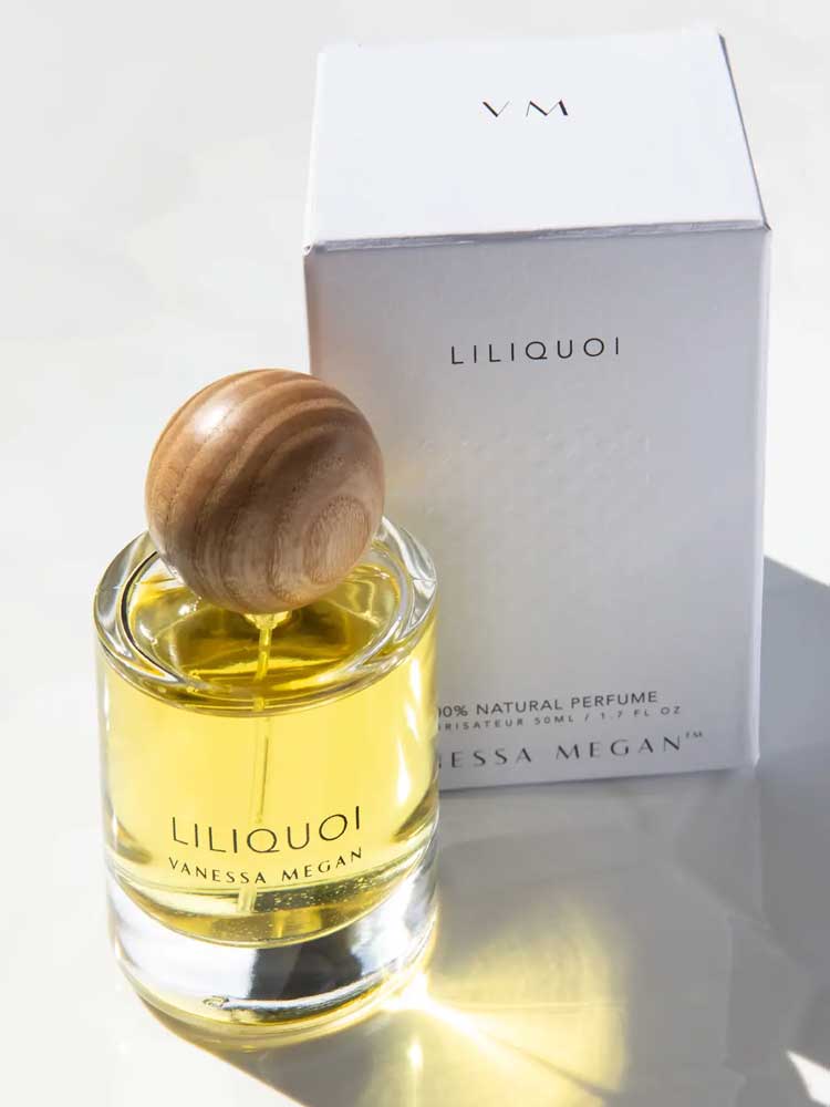 A glass bottle of Vanessa Megan Liliquoi 100% Natural Eau de Parfum 50ml with a round wooden cap sits before a white box marked Liliquoi and VM, sunlight highlighting this natural white-floral fragrance.