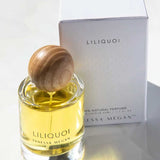A glass bottle of Vanessa Megan Liliquoi 100% Natural Eau de Parfum 50ml with a round wooden cap sits before a white box marked Liliquoi and VM, sunlight highlighting this natural white-floral fragrance.