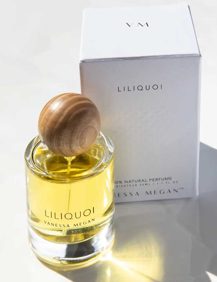 A glass bottle of Vanessa Megan Liliquoi 100% Natural Eau de Parfum 50ml with a round wooden cap sits before a white box marked Liliquoi and VM, sunlight highlighting this natural white-floral fragrance.