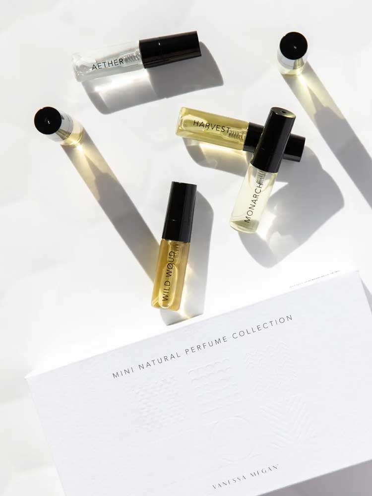 Six small Vanessa Megan 100% Natural Mini Eau de Parfum Collection vials (3ml each) with black caps are arranged on a white surface near the labeled box, their shadows cast in the sunlight.