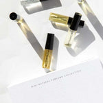 Six small Vanessa Megan 100% Natural Mini Eau de Parfum Collection vials (3ml each) with black caps are arranged on a white surface near the labeled box, their shadows cast in the sunlight.