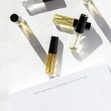 Six small Vanessa Megan 100% Natural Mini Eau de Parfum Collection vials (3ml each) with black caps are arranged on a white surface near the labeled box, their shadows cast in the sunlight.