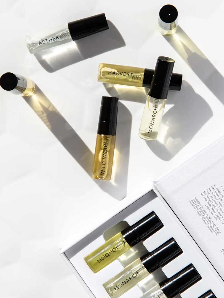 A flat lay of Vanessa Megan 100% Natural Mini Eau de Parfum Collection (6 x 3ml) bottles—AETHER, HARVEST, WILD INDIGO, MONARCH, and LIQUOR—shown beside the open box with more plant-based fragrance vials and a booklet.