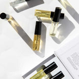 A flat lay of Vanessa Megan 100% Natural Mini Eau de Parfum Collection (6 x 3ml) bottles—AETHER, HARVEST, WILD INDIGO, MONARCH, and LIQUOR—shown beside the open box with more plant-based fragrance vials and a booklet.