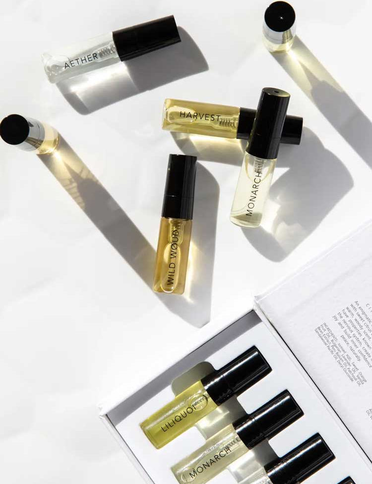 A flat lay of Vanessa Megan 100% Natural Mini Eau de Parfum Collection (6 x 3ml) bottles, AETHER, HARVEST, WILD INDIGO, MONARCH, and LIQUOR, shown beside the open box with more plant-based fragrance vials and a booklet.