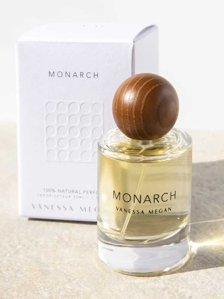 A bottle of Vanessa Megan Monarch 100% Natural Eau de Parfum 50ml with a round wooden cap sits in front of its white box on a light surface, illuminated by sunlight.