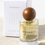 A bottle of Vanessa Megan Monarch 100% Natural Eau de Parfum 50ml with a round wooden cap sits in front of its white box on a light surface, illuminated by sunlight.