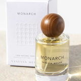A bottle of Vanessa Megan Monarch 100% Natural Eau de Parfum 50ml with a round wooden cap sits in front of its white box on a light surface, illuminated by sunlight.