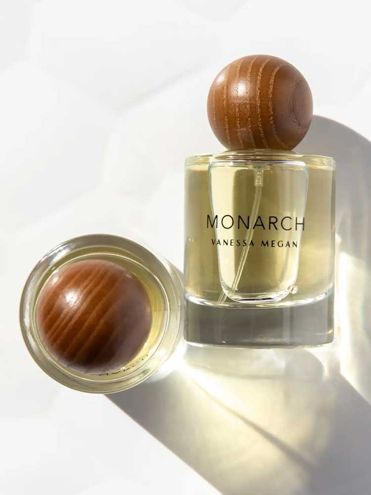 A clear glass bottle of Vanessa Megan Monarch 100% Natural Eau de Parfum 50ml with a round wooden cap sits upright beside another capped bottle on its side, both casting soft shadows—a refined display of natural perfumery.