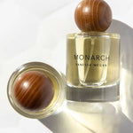 A clear glass bottle of Vanessa Megan Monarch 100% Natural Eau de Parfum 50ml with a round wooden cap sits upright beside another capped bottle on its side, both casting soft shadows—a refined display of natural perfumery.