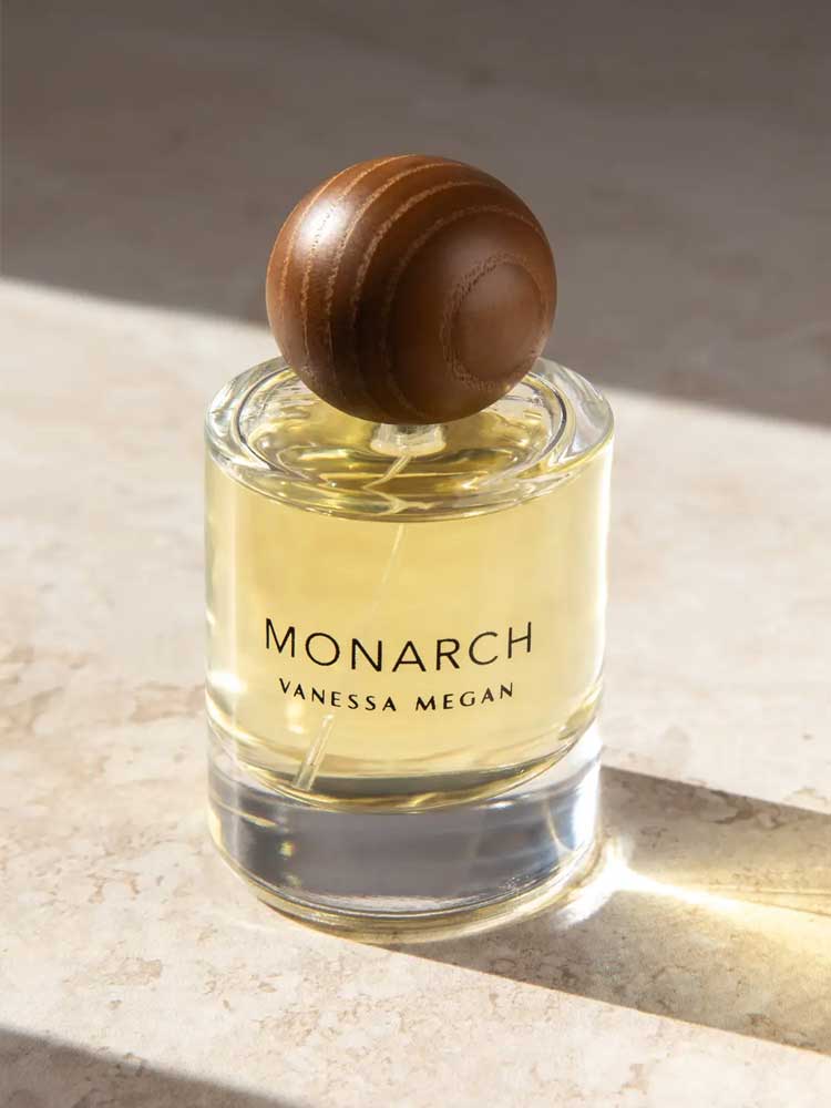 A glass bottle of Vanessa Megan Monarch 100% Natural Eau de Parfum 50ml, filled with yellow liquid and topped with a round brown wooden cap, rests on a sunlit stone surface.