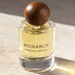 A glass bottle of Vanessa Megan Monarch 100% Natural Eau de Parfum 50ml, filled with yellow liquid and topped with a round brown wooden cap, rests on a sunlit stone surface.