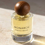 A glass bottle of Vanessa Megan Monarch 100% Natural Eau de Parfum 50ml, filled with yellow liquid and topped with a round brown wooden cap, rests on a sunlit stone surface.