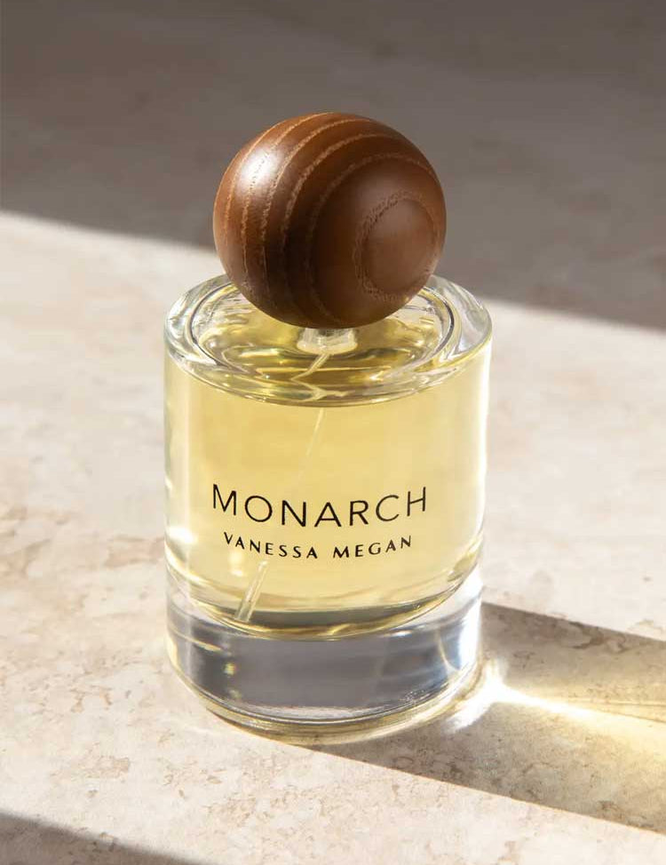 A glass bottle of Vanessa Megan Monarch 100% Natural Eau de Parfum 50ml, filled with yellow liquid and topped with a round brown wooden cap, rests on a sunlit stone surface.