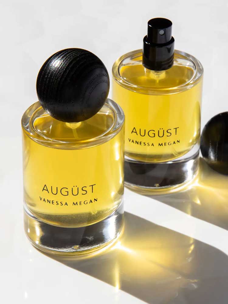 Two bottles of Vanessa Megan Augüst 100% Natural Eau de Parfum 50ml sit on a light surface—one capped, one with a spray nozzle. Their yellow glass and soft shadows evoke the refined, botanical essence of the fragrance.