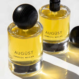 Two bottles of Vanessa Megan Augüst 100% Natural Eau de Parfum 50ml sit on a light surface—one capped, one with a spray nozzle. Their yellow glass and soft shadows evoke the refined, botanical essence of the fragrance.