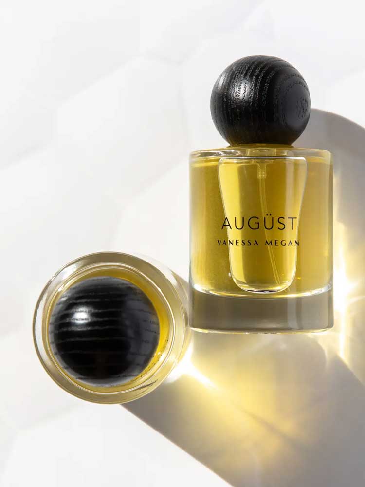 Two glass bottles of Vanessa Megan Augüst 100% Natural Eau de Parfum 50ml, featuring yellow liquid and round black textured caps—one upright, one lying on its side—evoke elegant botanical-inspired fragrance.