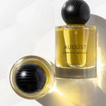 Two glass bottles of Vanessa Megan Augüst 100% Natural Eau de Parfum 50ml, featuring yellow liquid and round black textured caps—one upright, one lying on its side—evoke elegant botanical-inspired fragrance.