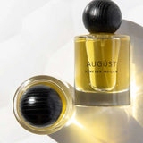 Two glass bottles of Vanessa Megan Augüst 100% Natural Eau de Parfum 50ml, featuring yellow liquid and round black textured caps—one upright, one lying on its side—evoke elegant botanical-inspired fragrance.