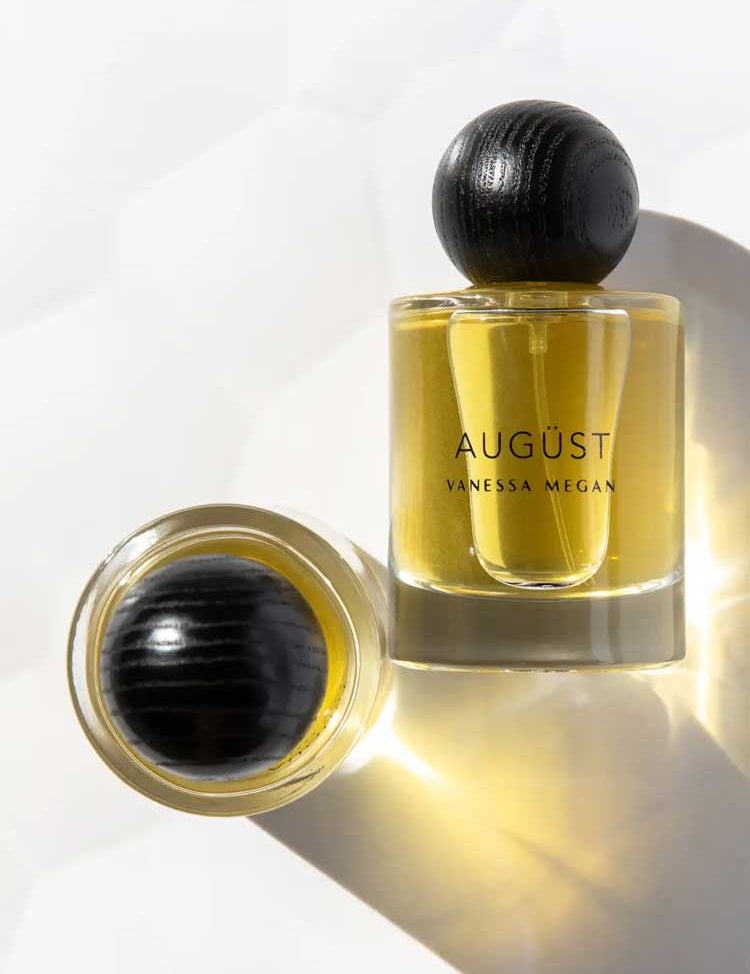 Two glass bottles of Vanessa Megan Augüst 100% Natural Eau de Parfum 50ml, featuring yellow liquid and round black textured caps, one upright, one lying on its side, evoke elegant botanical-inspired fragrance.