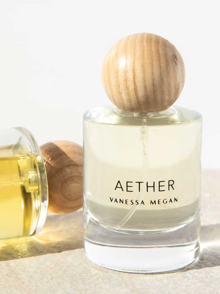 A clear glass bottle of Vanessa Megan Aether 100% Natural Eau de Parfum 50ml with a round wooden cap stands upright beside a similar bottle on its side, both filled with light yellow fragrance, handcrafted in Australia.