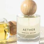 A clear glass bottle of Vanessa Megan Aether 100% Natural Eau de Parfum 50ml with a round wooden cap stands upright beside a similar bottle on its side, both filled with light yellow fragrance, handcrafted in Australia.