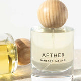 A clear glass bottle of Vanessa Megan Aether 100% Natural Eau de Parfum 50ml with a round wooden cap stands upright beside a similar bottle on its side, both filled with light yellow fragrance, handcrafted in Australia.