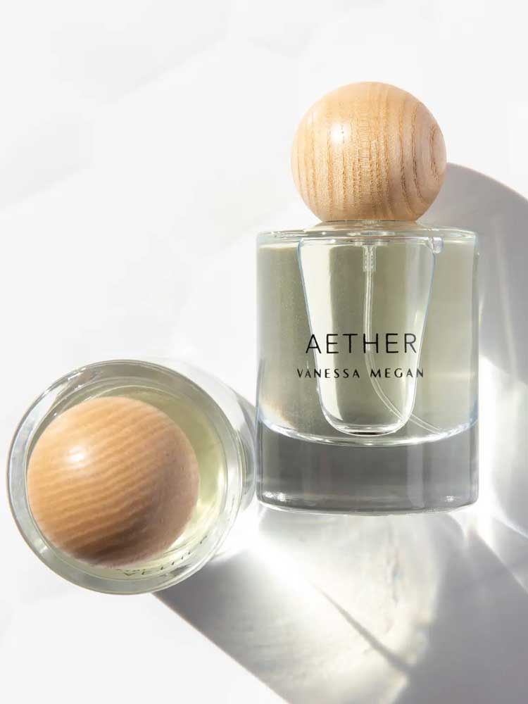A clear Vanessa Megan Aether 100% Natural Eau de Parfum 50ml stands upright with a round wooden cap, handcrafted in Australia with plant-derived ingredients. Another similar bottle lies on its side, casting soft shadows on a white surface.
