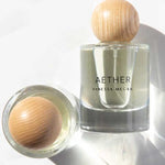 A clear Vanessa Megan Aether 100% Natural Eau de Parfum 50ml stands upright with a round wooden cap, handcrafted in Australia with plant-derived ingredients. Another similar bottle lies on its side, casting soft shadows on a white surface.