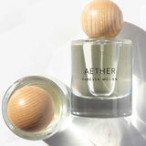 A clear Vanessa Megan Aether 100% Natural Eau de Parfum 50ml stands upright with a round wooden cap, handcrafted in Australia with plant-derived ingredients. Another similar bottle lies on its side, casting soft shadows on a white surface.