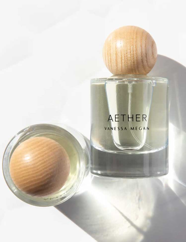 A clear Vanessa Megan Aether 100% Natural Eau de Parfum 50ml stands upright with a round wooden cap, handcrafted in Australia with plant-derived ingredients. Another similar bottle lies on its side, casting soft shadows on a white surface.