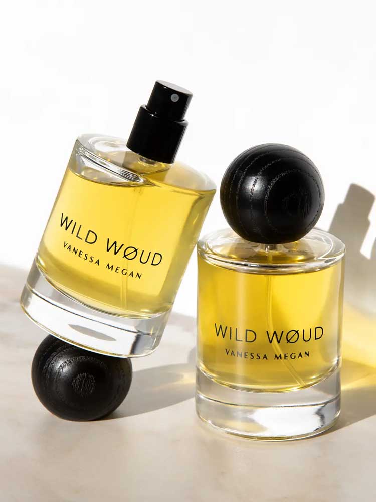 Two bottles of Vanessa Megan Wild Wøud 100% Natural Eau de Parfum 50ml, both with black caps and yellow liquid, sit on a white surface with soft shadows—one upright, one tilted.