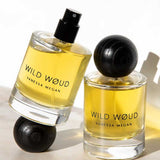 Two bottles of Vanessa Megan Wild Wøud 100% Natural Eau de Parfum 50ml, both with black caps and yellow liquid, sit on a white surface with soft shadows—one upright, one tilted.