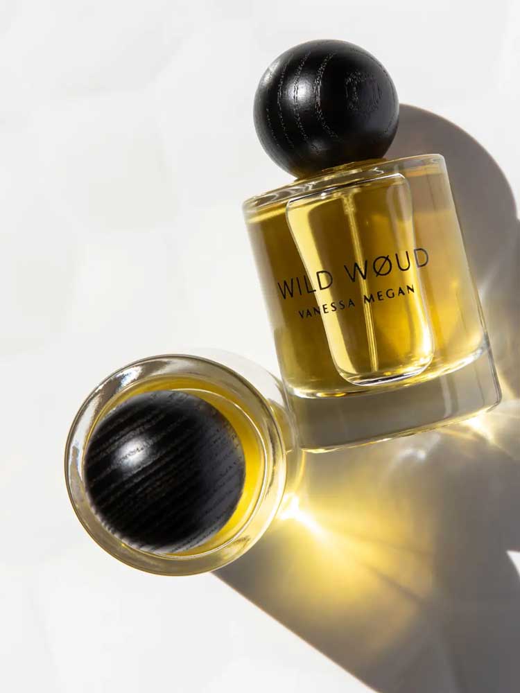 Two bottles of Vanessa Megan Wild Wøud 100% Natural Eau de Parfum 50ml, with golden liquid and black rounded caps, rest on a white surface—one upright and one lying down—casting soft shadows.