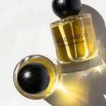 Two bottles of Vanessa Megan Wild Wøud 100% Natural Eau de Parfum 50ml, with golden liquid and black rounded caps, rest on a white surface—one upright and one lying down—casting soft shadows.